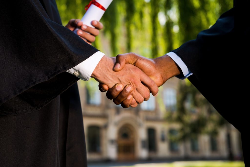 Congratulations! Close-up of two men in graduation gowns holding diplomas and shaking hands
